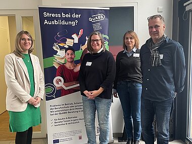 Referentin und drei Teilnehmende des Thementags stehen vor einem Roll-Up von QuABB