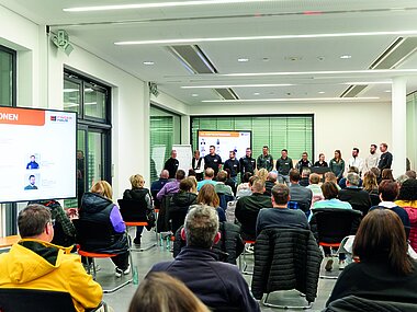 Foto der Verastaltung für Azubi-Eltern: Personen sitzen in einem Raum auf Stühlen, vorne stellen sich die Ausbildungsverantworlichen vor.
