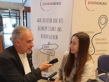 Ein Mann und eine Frau unterhalten sich. Der Mann hat ein Mikrofon in der Hand und hält es der Frau hin.