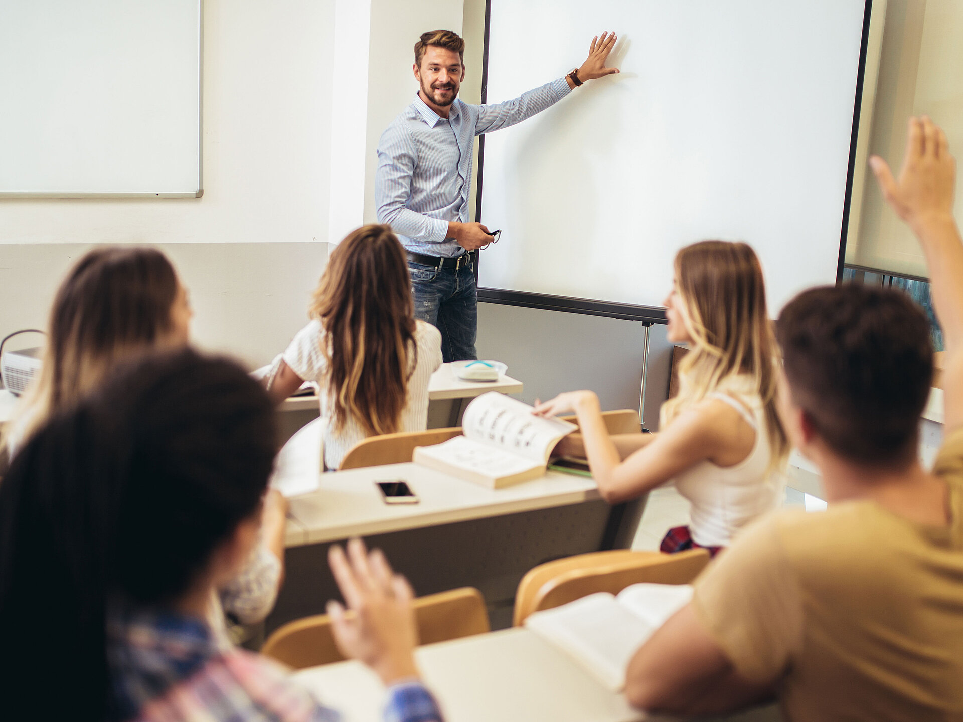 Junger Lehrer in einem hellblauen Hemd vor einer weißen Tafel in einer Schulklasse mit Schülerinnen und Schülern, die sich melden.