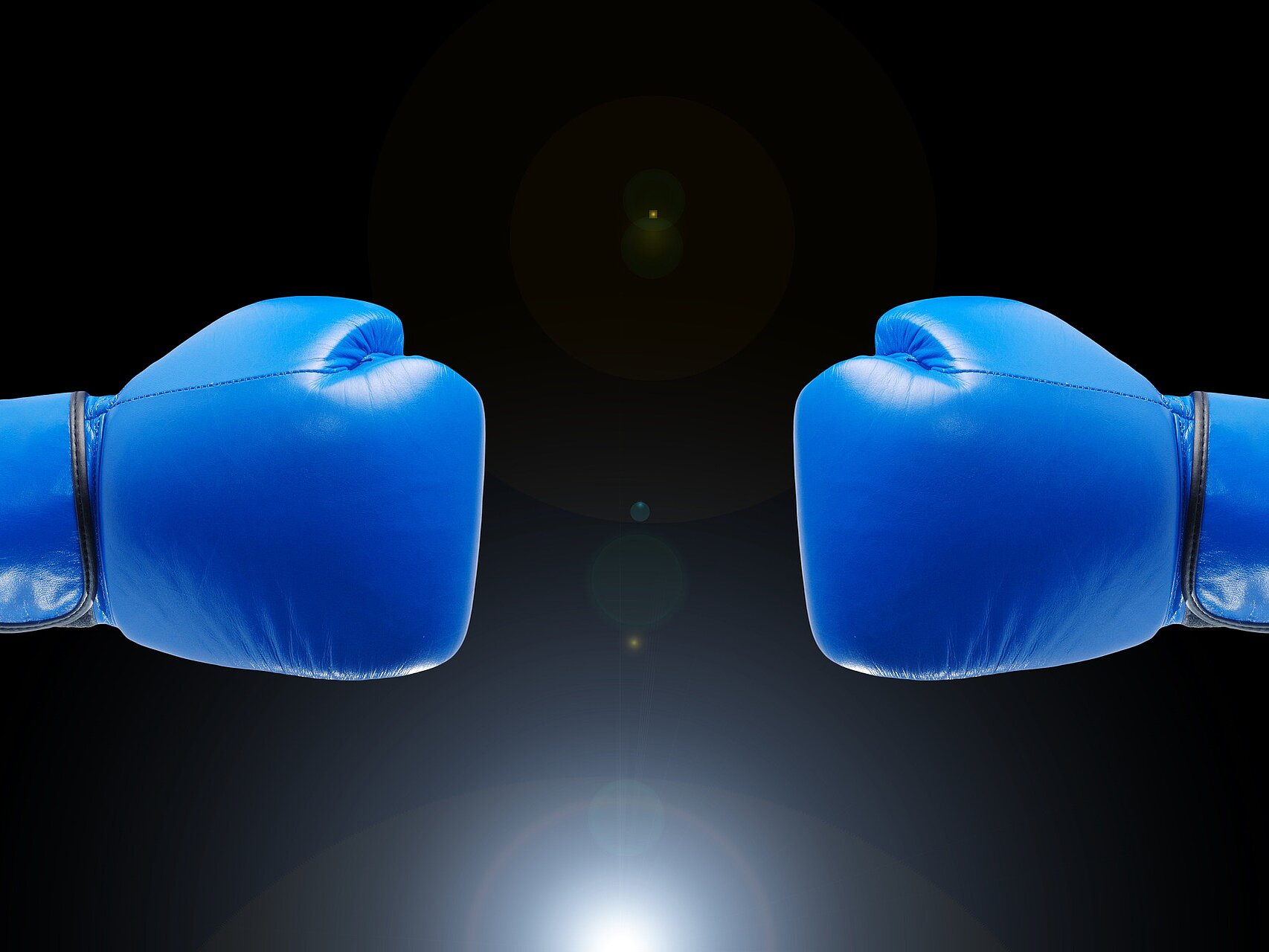 Zwei blaue gegeneinander gerichtete Boxhandschuhe vor einem dunklen Hintergrund.