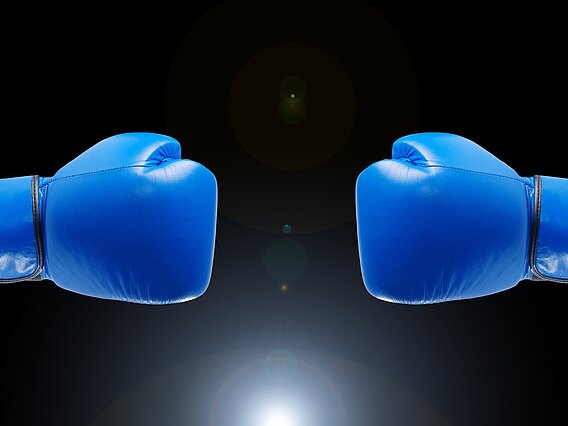 Zwei blaue gegeneinander gerichtete Boxhandschuhe vor einem dunklen Hintergrund.